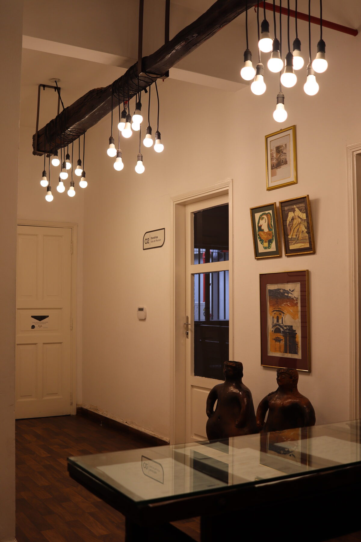 Salón de la sede con lámparas colgantes, mesa de vidrio, cuadros y dos figuras de cerámica al fondo